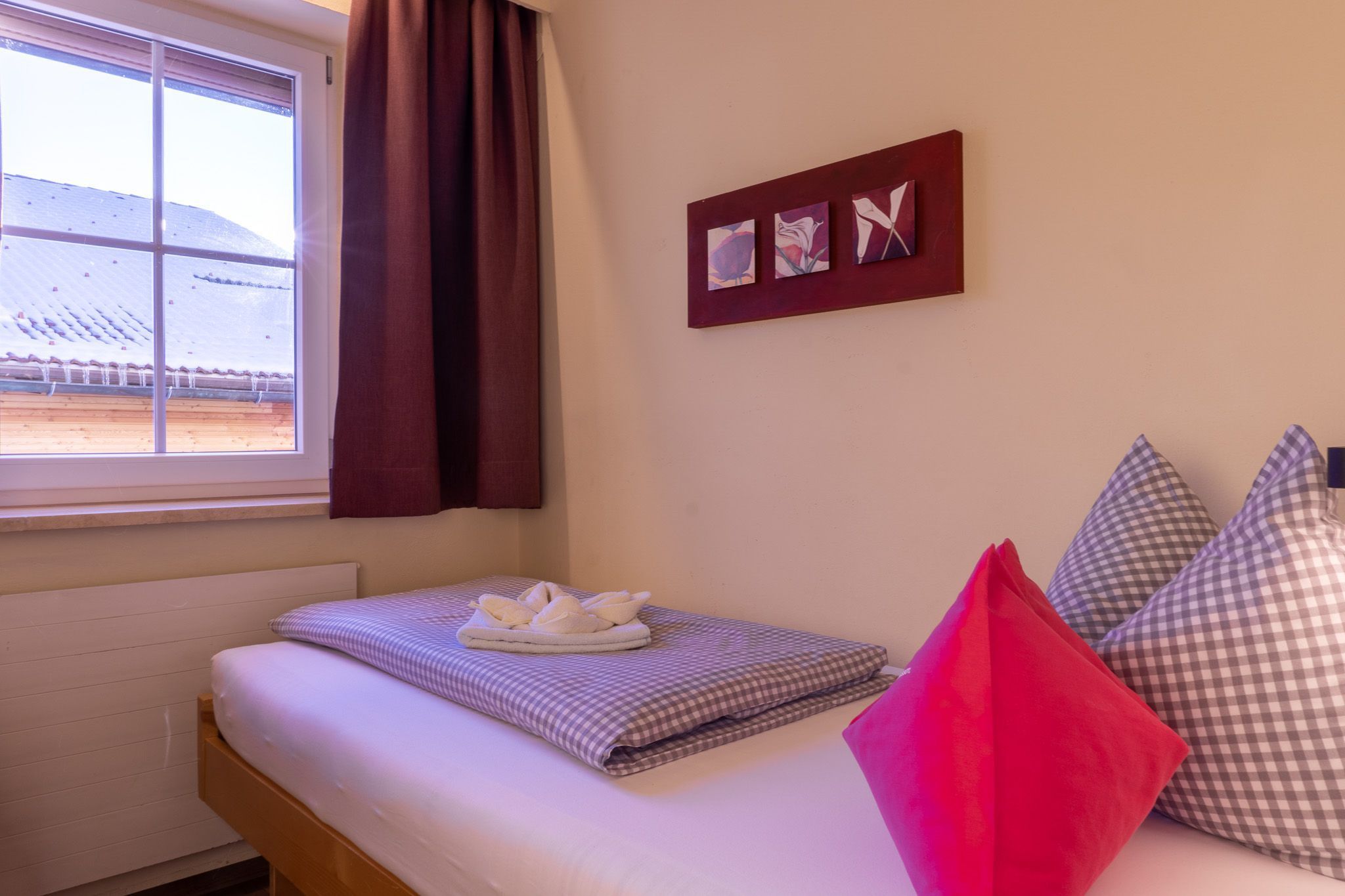 A neatly made bed with a checkered blanket, a pillow, and a folded towel. There is a window with curtains, a wall-mounted picture frame, and a radiator.