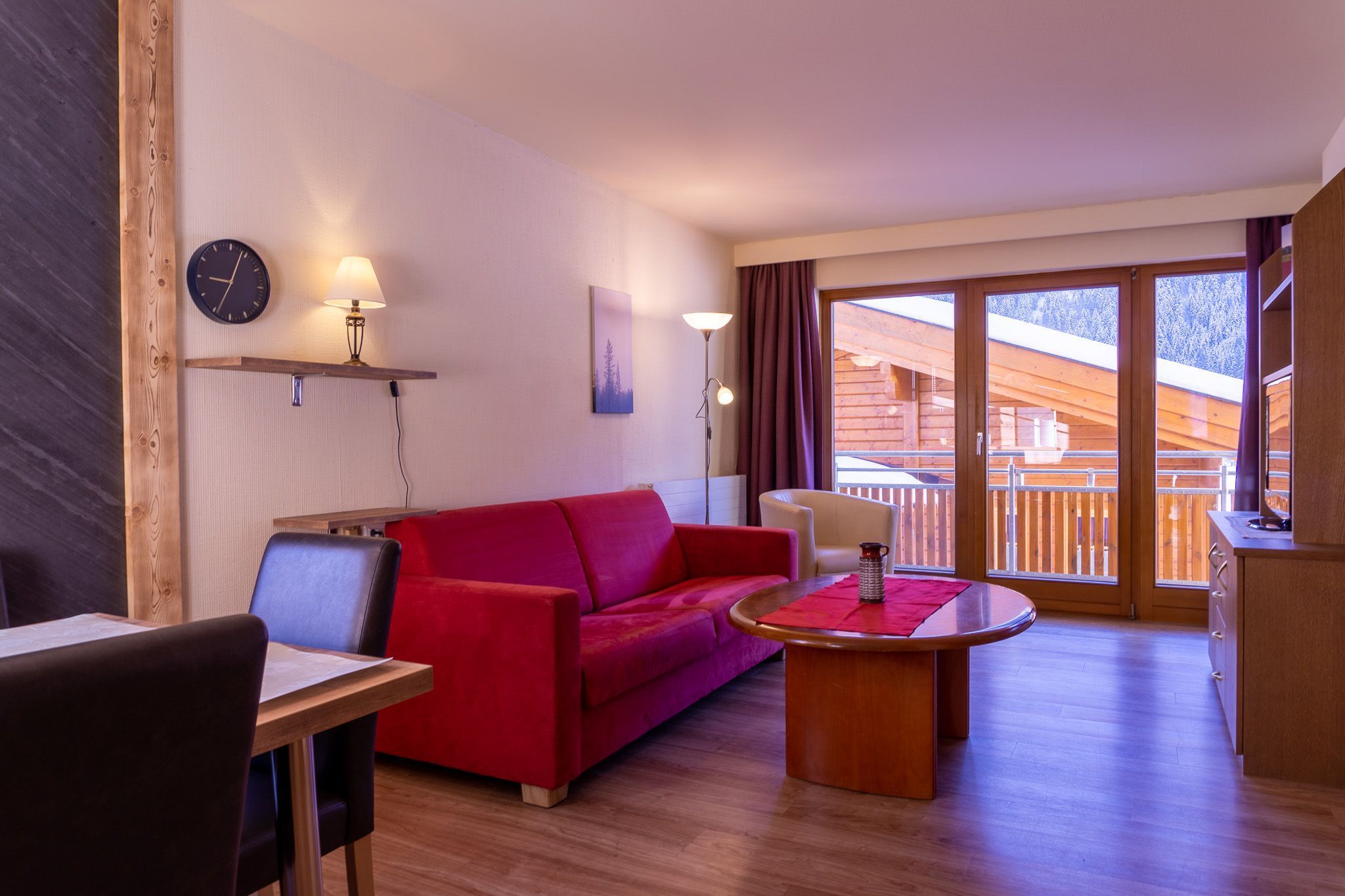 Una habitación acogedora con un sofá rojo, una mesa de madera, una silla, un reloj y una lámpara en un estante.