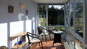 A sunlit patio with a table and chairs, a small shelf with a bowl, and a view of greenery through large windows. - Apartment 01 in the fisherman's house with terrace - Apartment Fischers Fritze with winter garden and (Sellin)