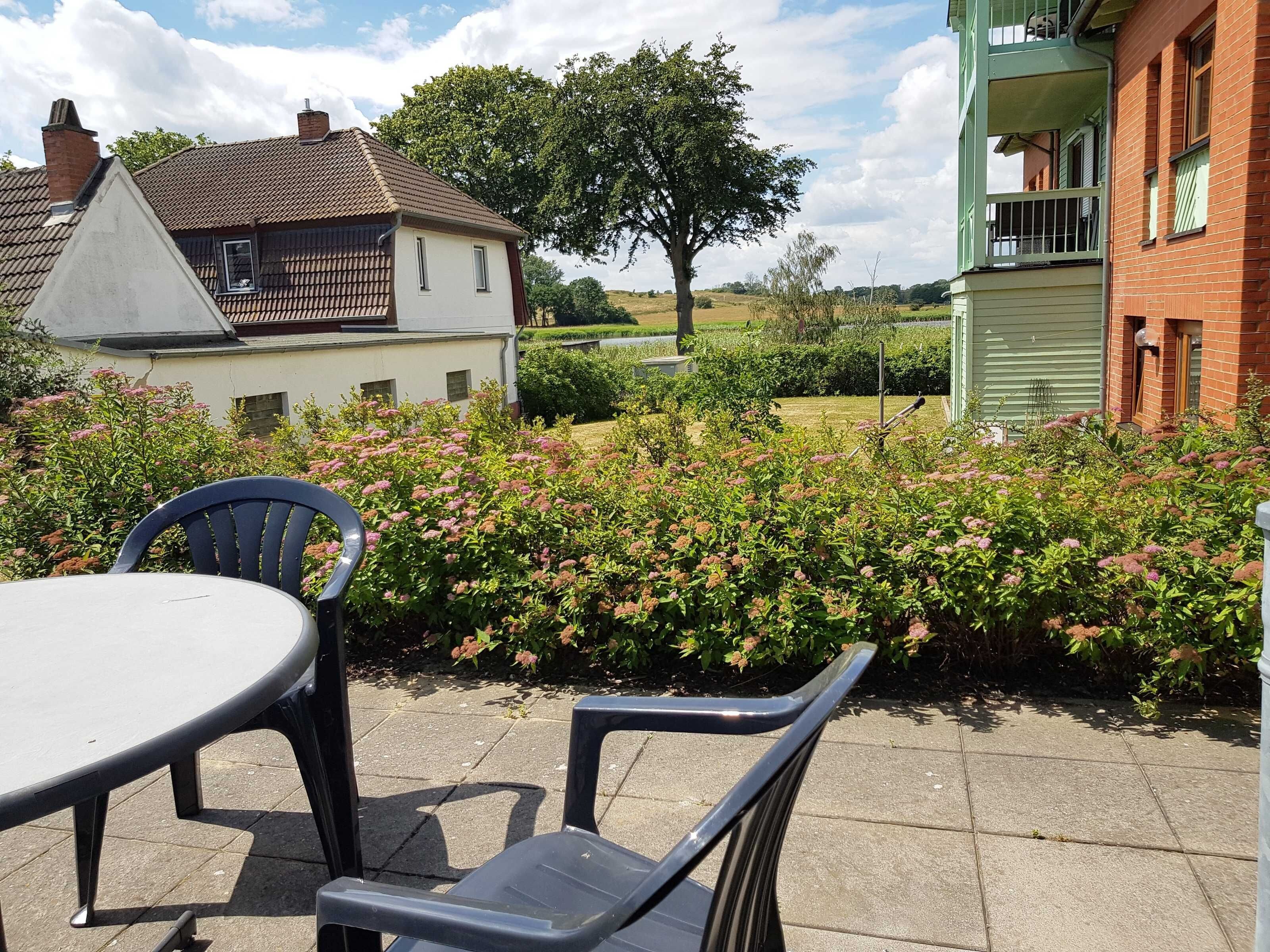 A patio with a table and chairs, surrounded by flowering bushes and a view of a residential area with houses and a tree.
