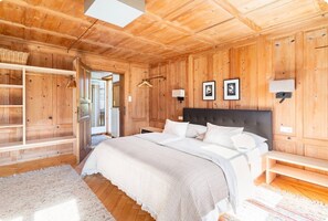 A wooden-paneled bedroom with a bed, bedside tables, a hanging light, and a view through an open door.