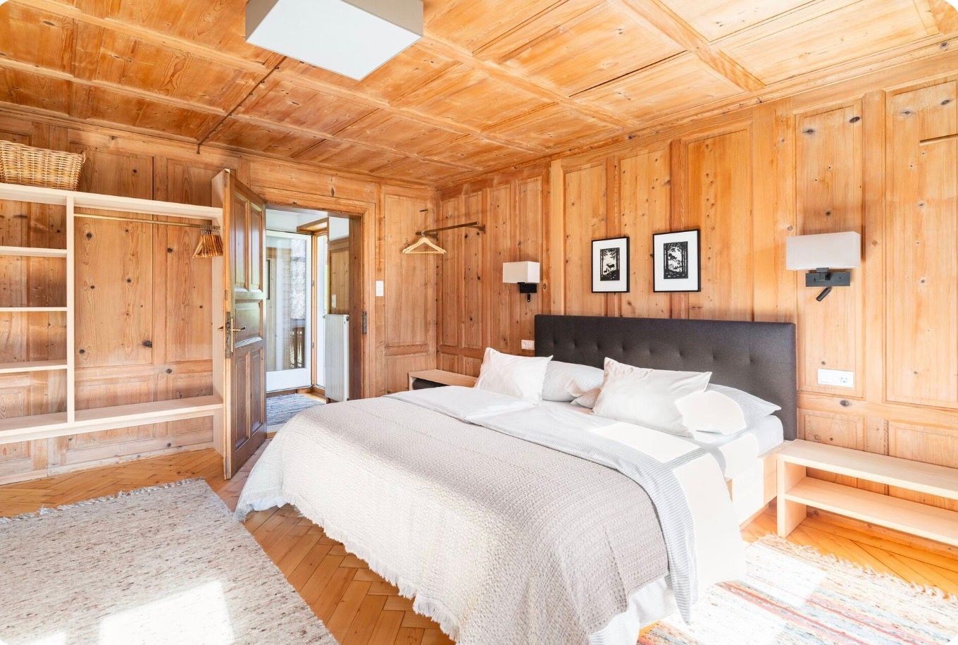 A wooden-paneled bedroom with a bed, bedside tables, a hanging light, and a view through an open door.