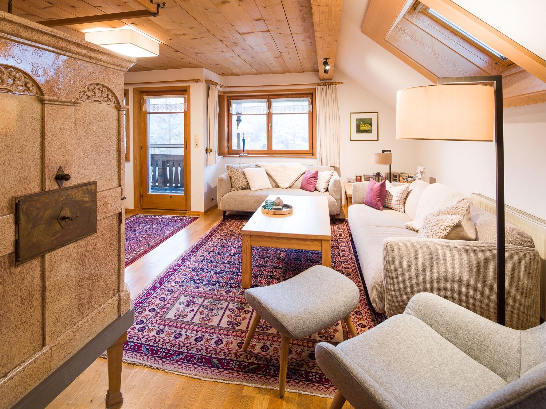 A cozy living room with a sofa, armchair, and a coffee table. There is a patterned rug on the floor, a door with a handle, and a window with curtains.