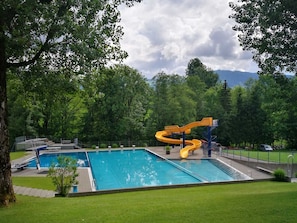 A swimming pool with a water slide, surrounded by trees and a cloudy sky.
