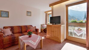 Ein Wohnzimmer mit einer Couch, einem Fernseher auf einer Holzeinrichtung, einem Tisch mit kariertem Tuch und einem Balkon mit Bergblick.