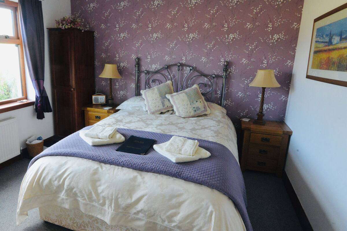 A neatly made bed with a purple blanket, two pillows, and two folded towels. There are two bedside tables with lamps and a small radio. The room has purple floral wallpaper and a window with curtains.