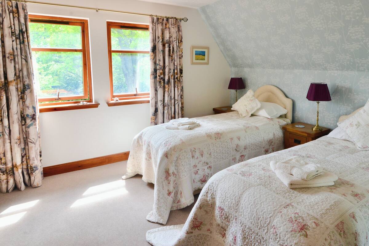 A bedroom with two beds, floral bedding, a nightstand with lamps, and a window with curtains.