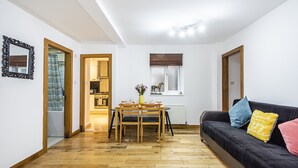 Un'ampia zona giorno con pavimento in legno, un divano nero con cuscini colorati, un tavolo da pranzo con sedie e una piccola cucina visibile attraverso una porta aperta.