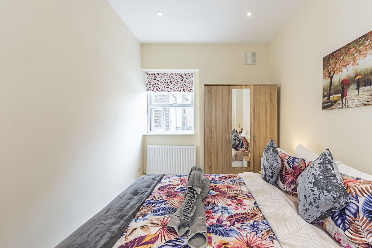 A bedroom with a bed, a window with blinds, a radiator, a wooden wardrobe, and a painting on the wall.