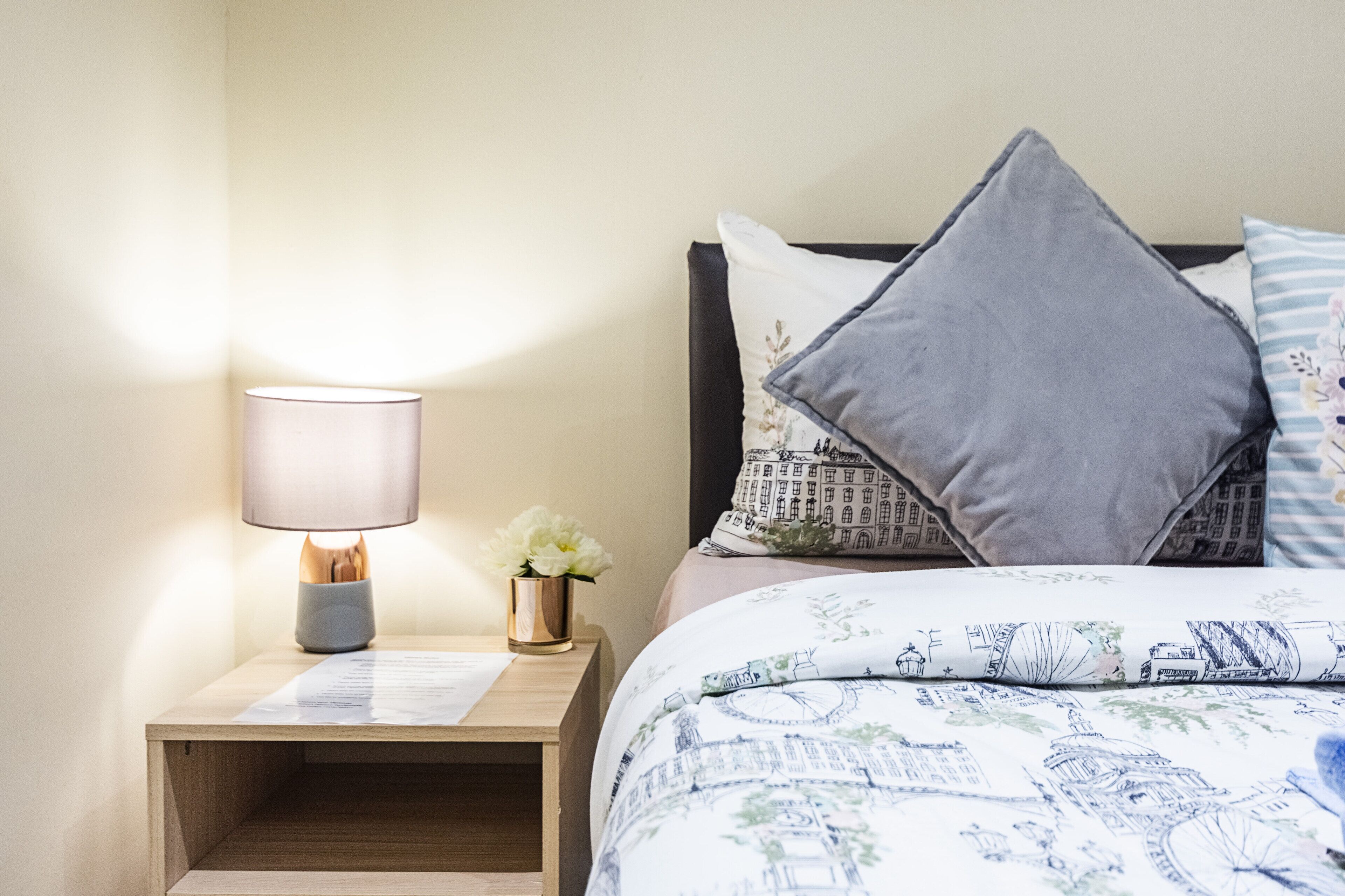 Una cama bien tendida con ropa de cama estampada, una lámpara de noche, un pequeño jarrón con flores y una mesita de noche de madera.