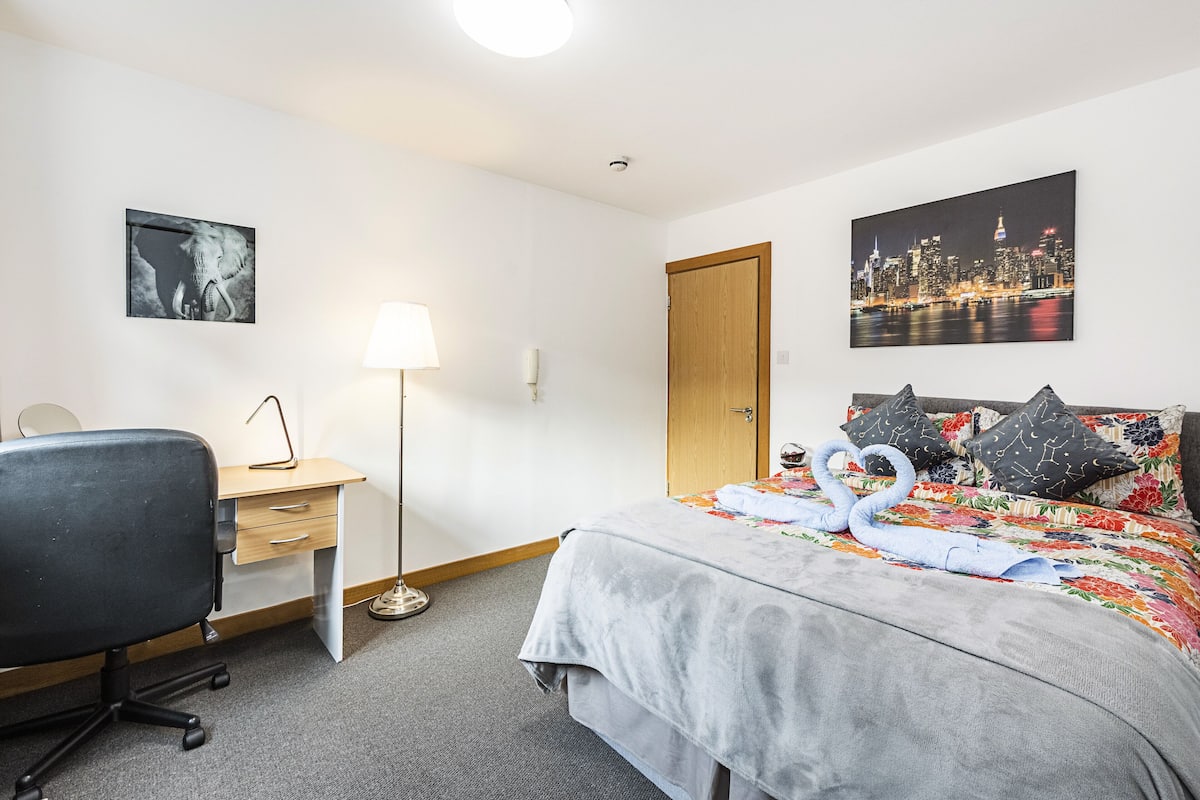 A hotel room with a bed, a desk, a chair, a lamp, and two wall-mounted pictures.