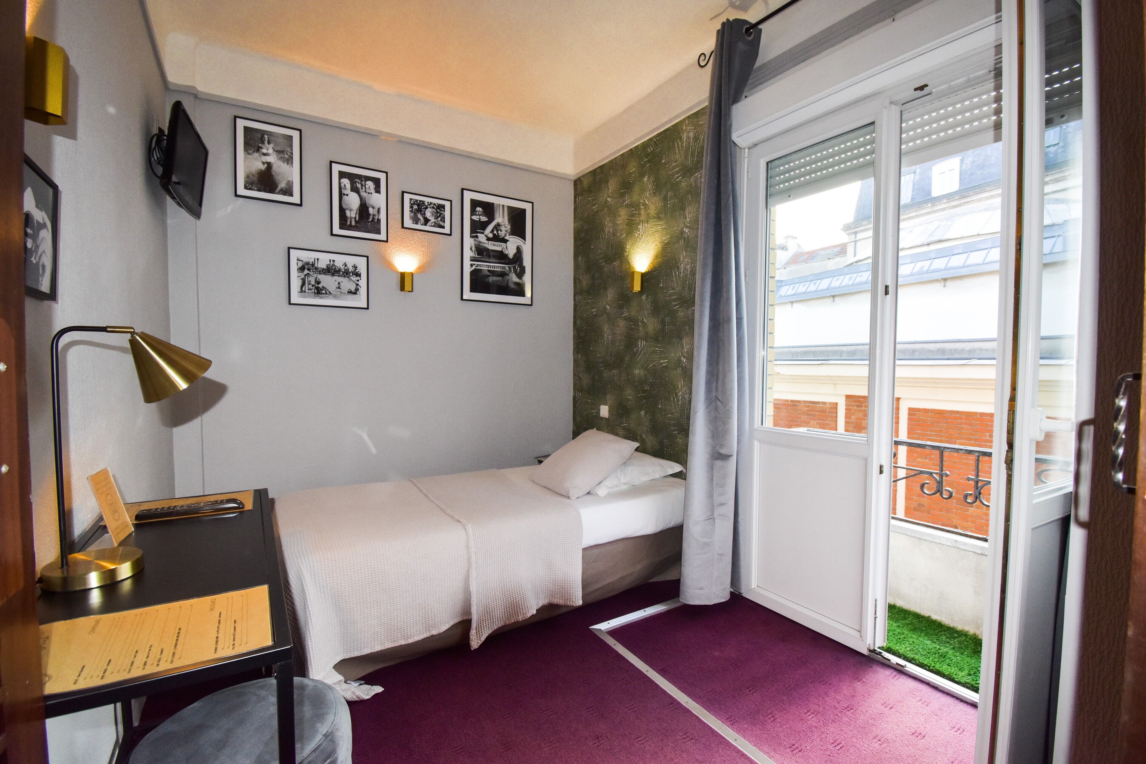 A hotel room with a bed, a desk with a lamp, a window with curtains, and a balcony with a view of buildings.