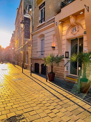A cobblestone street with a hotel on the right, featuring a large potted plant and a sign with a clock.