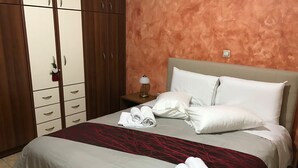 Una camera d'albergo con un letto, cuscini bianchi, una coperta grigio-rossa, un comodino con una lampada e un armadio con diversi cassetti.