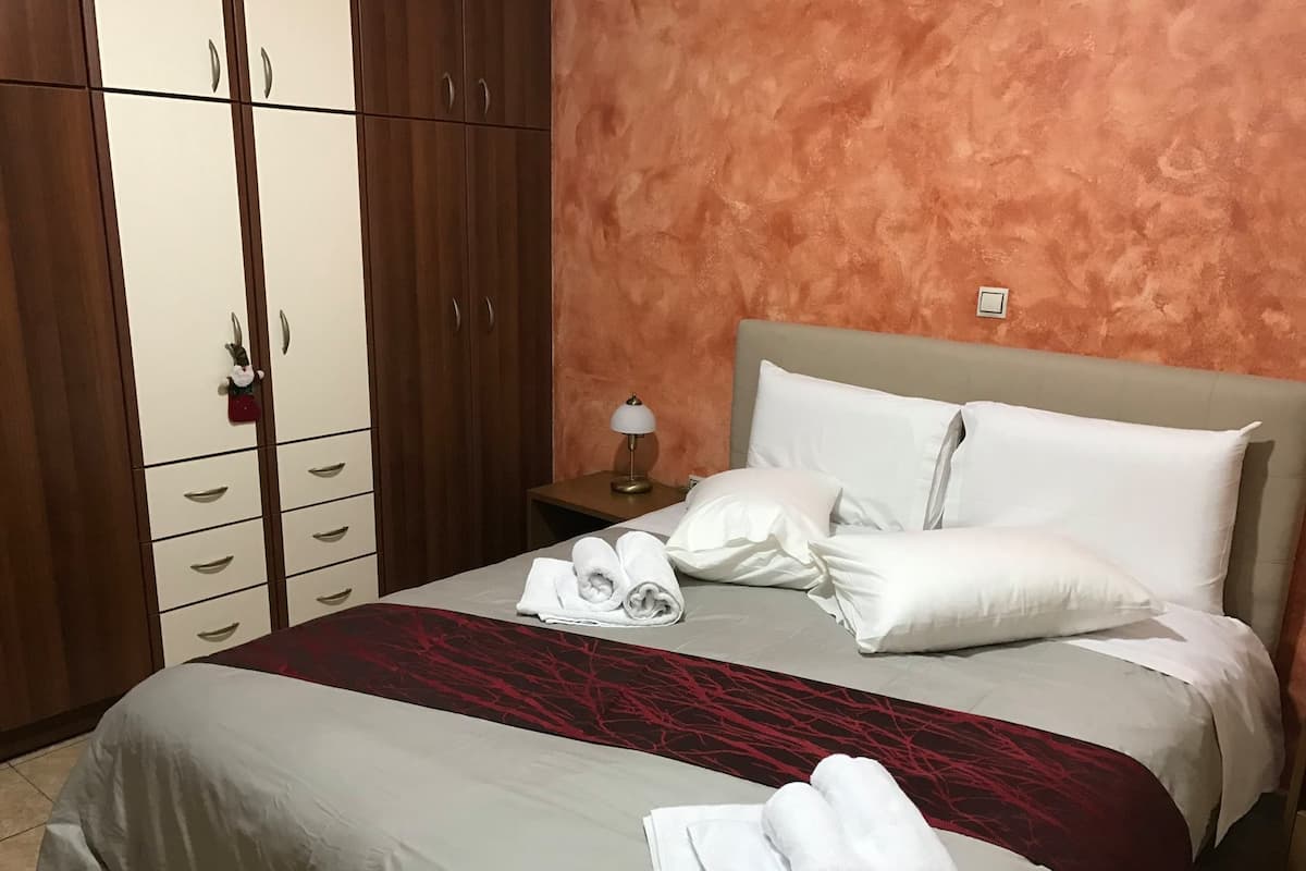 A hotel room with a bed, white pillows, a red and grey bedspread, a nightstand with a lamp, and a wardrobe with multiple drawers.