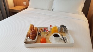Una cama bien hecha con ropa blanca, una bandeja con cruasanes, jugo de naranja, café y un tazón pequeño de cereales.