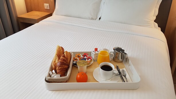Una cama bien hecha con ropa blanca, una bandeja con cruasanes, jugo de naranja, café y un tazón pequeño de cereales.