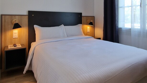 Una cama bien hecha con sábanas blancas, dos lámparas de noche y una ventana con cortinas.