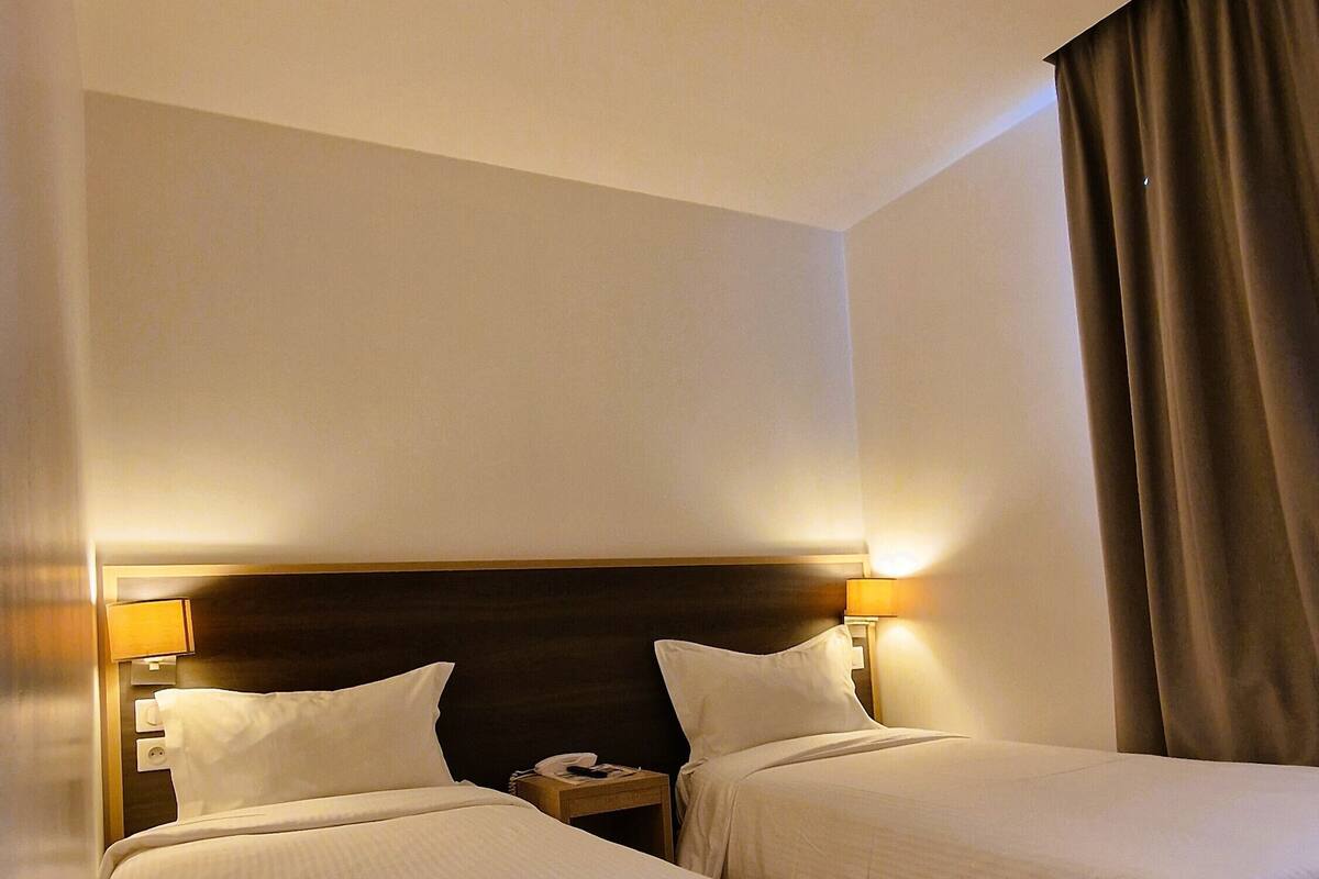 A hotel room with two single beds, a nightstand with a lamp, and a window with curtains.