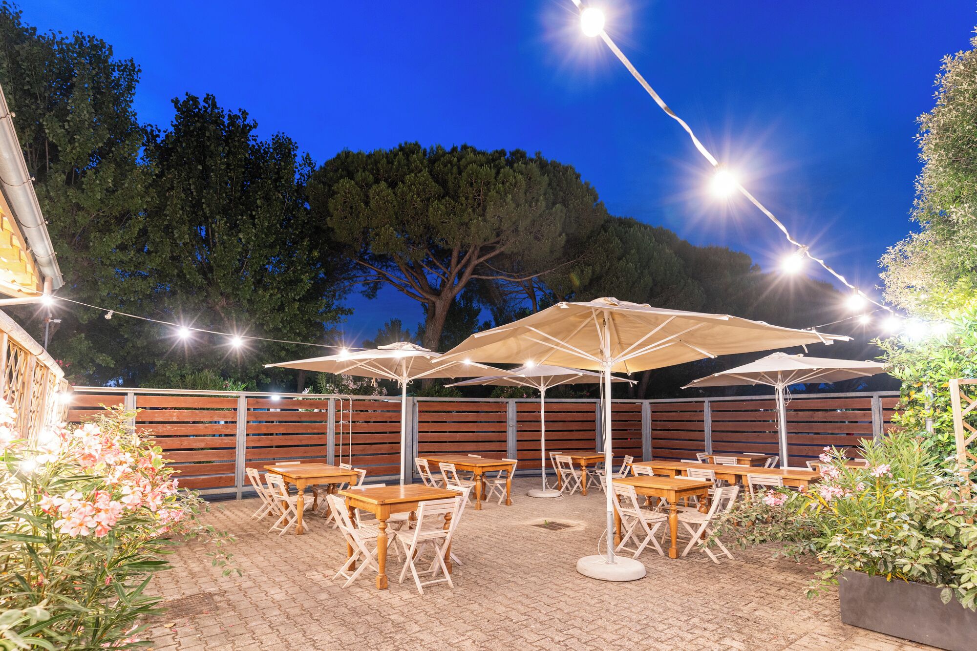 An outdoor patio with tables and umbrellas, surrounded by a wooden fence and trees.