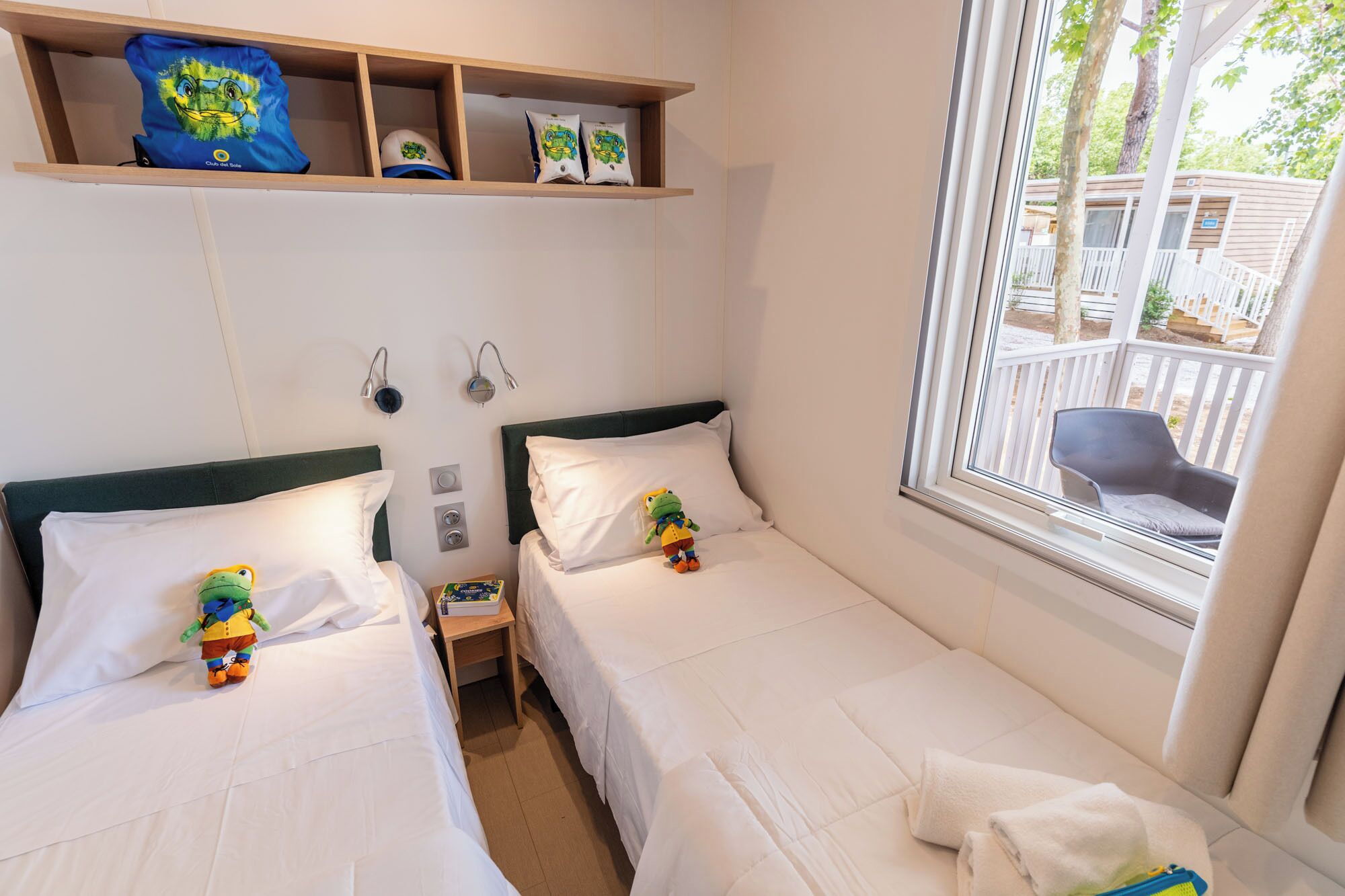 A hotel room with two beds, a shelf with hats, a window with a view, and a small table with a toy.