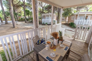 Un área de estar al aire libre cubierta, con una mesa dispuesta para el desayuno, rodeada de árboles y casas móviles.