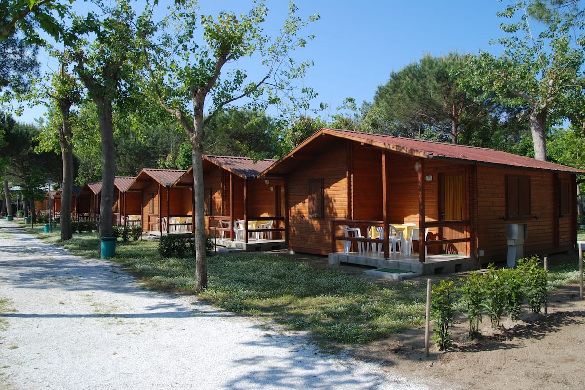 A row of wooden cabins with outdoor seating, surrounded by trees and a gravel path.