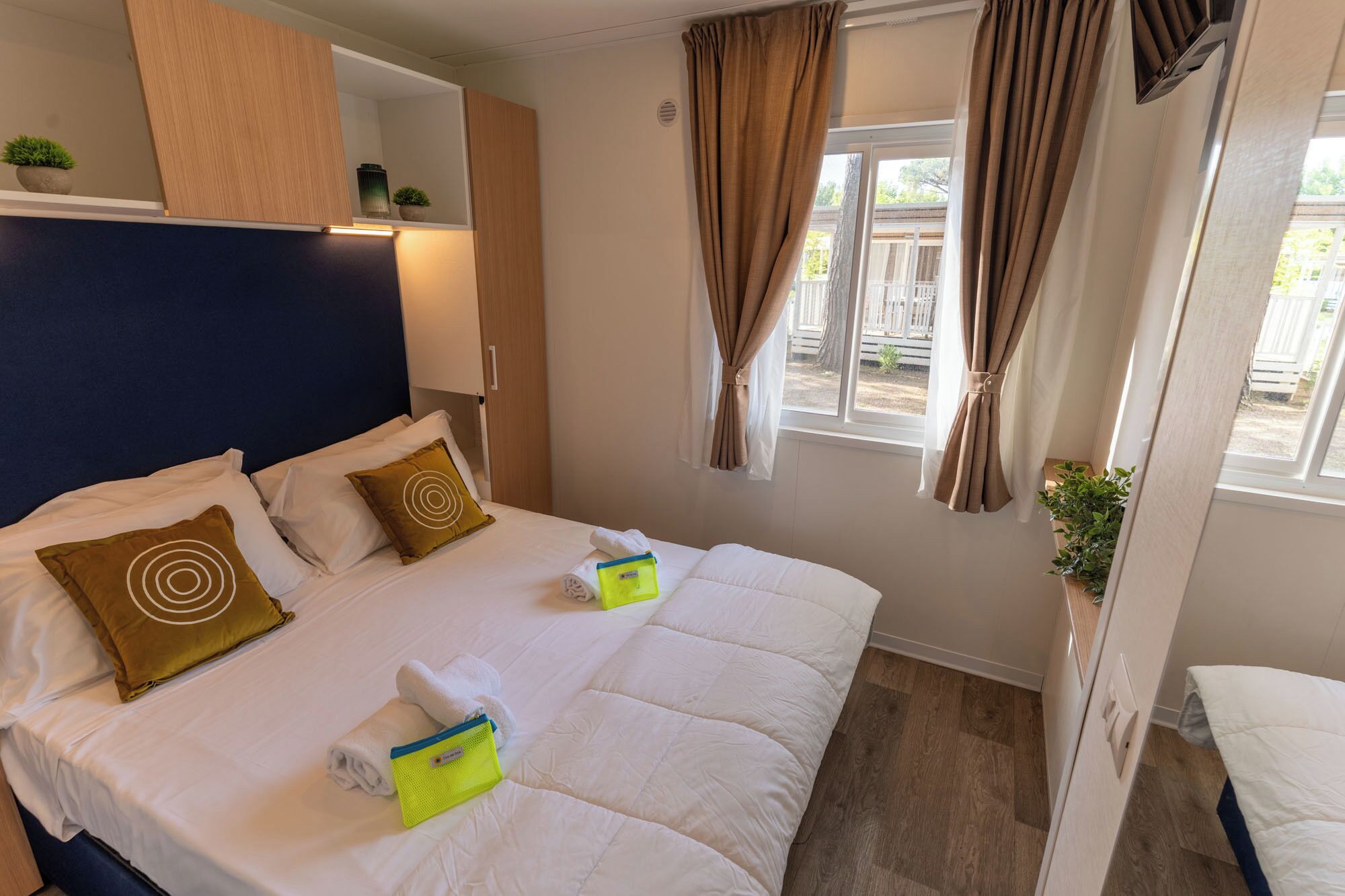 A neatly made bed with pillows, a bedside table with a lamp, a window with curtains, and a small plant on the windowsill.