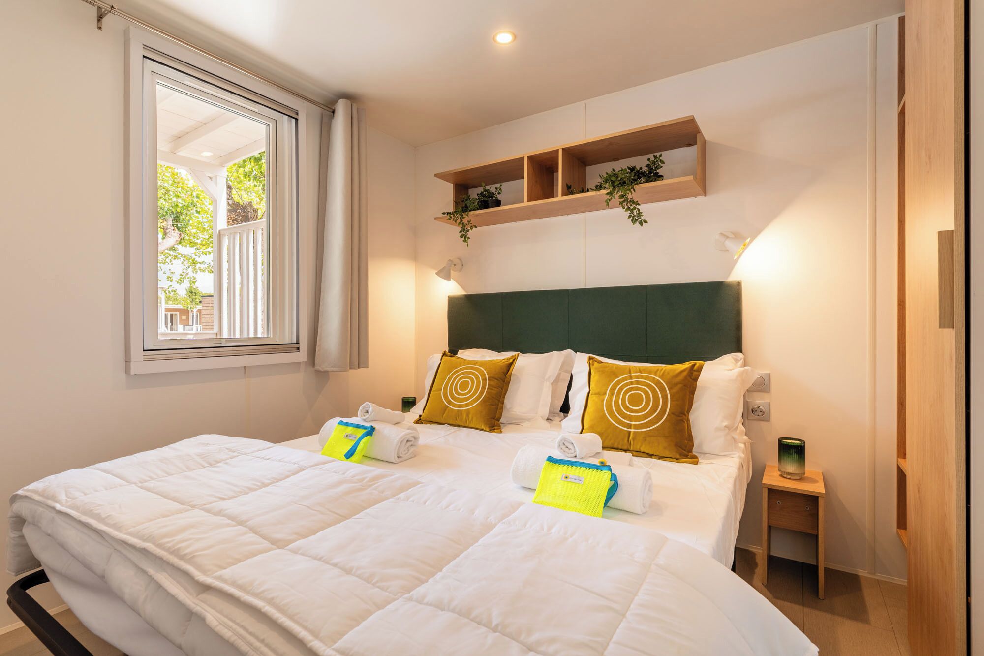 A hotel room with a large bed, a window with curtains, a bedside table, and a shelf with plants.
