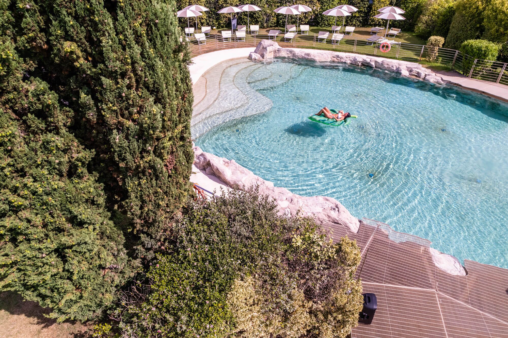 Une piscine avec une personne sur un flotteur, entourée d’arbres et de chaises longues.