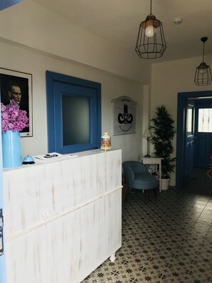 Un espace d’accueil avec un comptoir blanc, une fenêtre à cadre bleu, une affiche, une plante en pot et une chaise bleue.