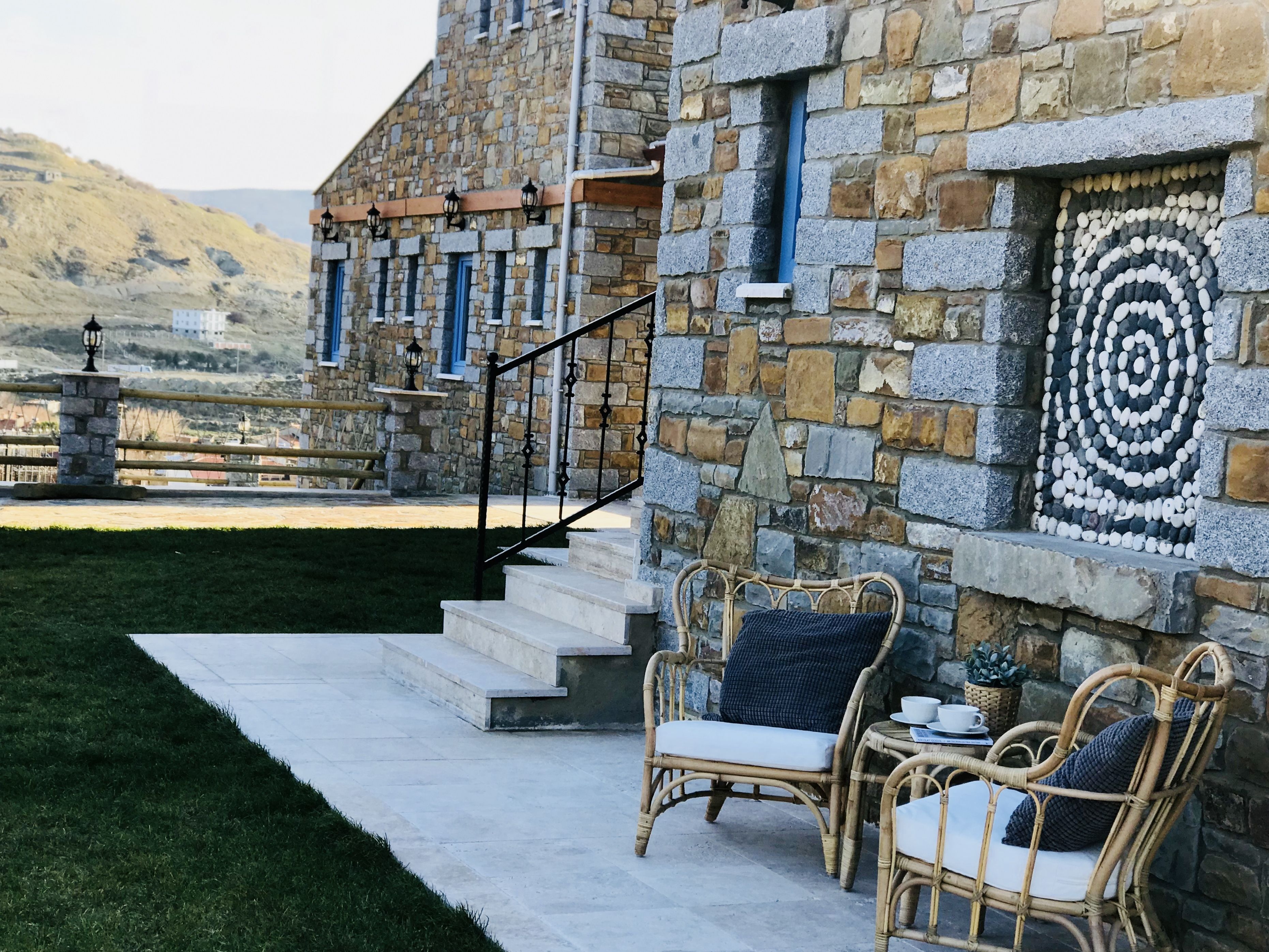 A stone building with a stone staircase leading to a balcony. There are wicker chairs and a table with cups and saucers on the balcony.
