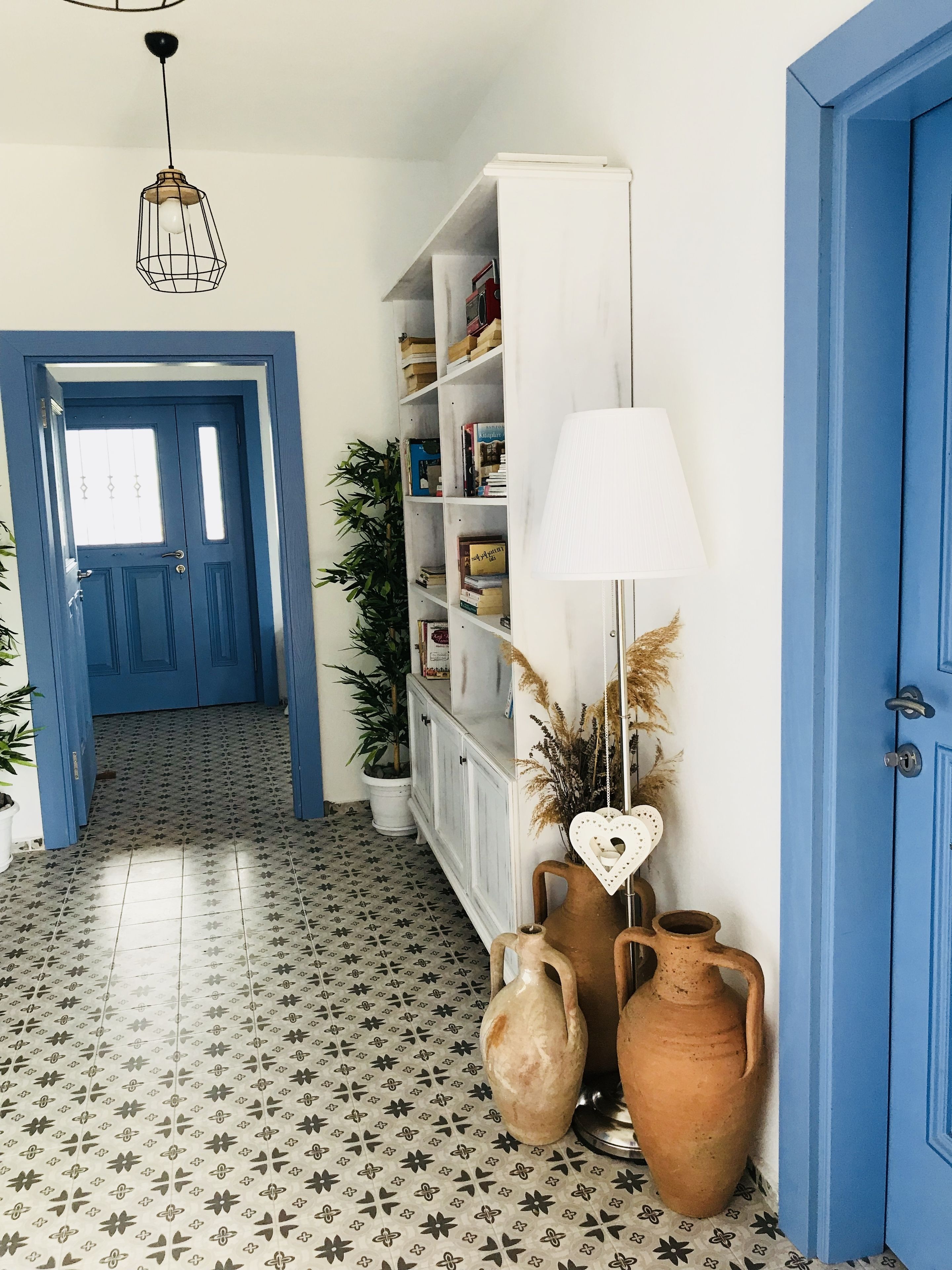 A hallway with patterned floor tiles, a white bookshelf, a blue door, and a hanging light fixture.