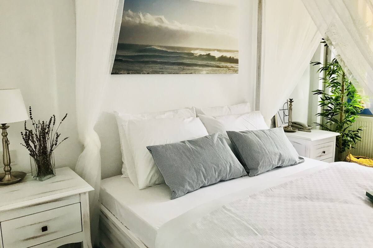 A bedroom with a bed, white nightstands, a lamp, a vase with flowers, and a window with a view of the sea.