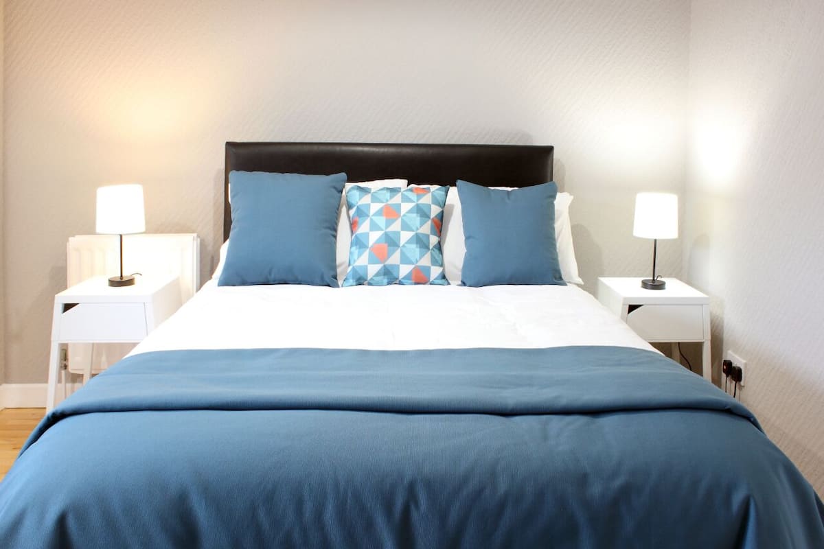 A neatly made bed with a blue bedspread and pillows, flanked by white nightstands with lamps.