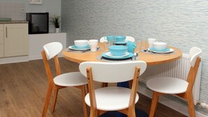 A round wooden dining table with four white chairs, blue and white tableware, and a kitchen area with cabinets and a microwave. - KC Accommodation (Dungannon)