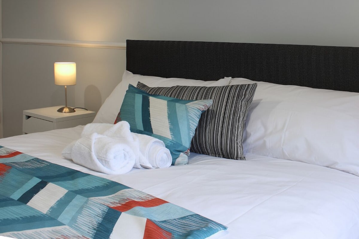 A neatly made bed with white linens, a teal and red patterned blanket, and rolled white towels.
