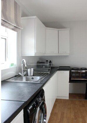 A compact kitchen with white cabinets, a stainless steel sink, a dishwasher, and a wooden floor. - KC Accommodation (Dungannon)