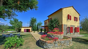 Une maison à deux étages, avec des fondations en pierre et des volets rouges, entourée d’un jardin bien entretenu, agrémenté de plantes fleuries et d’une pelouse.