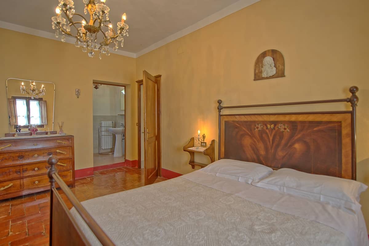 Een slaapkamer met een houten bed, een dressoir, een kroonluchter en een zichtbare badkamer door een open deur.