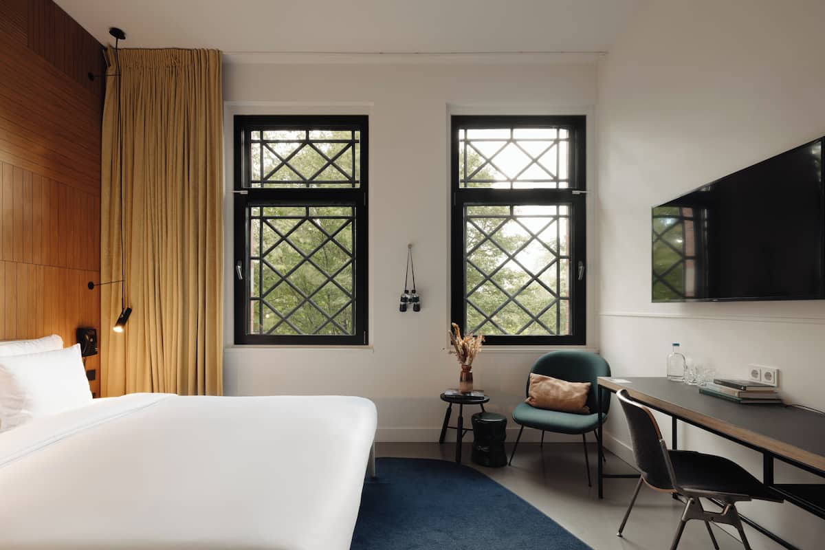 Habitación de hotel con una cama, dos ventanas con marcos negros, un escritorio con una silla, un televisor montado en la pared y una pequeña mesa con un jarrón y una botella.