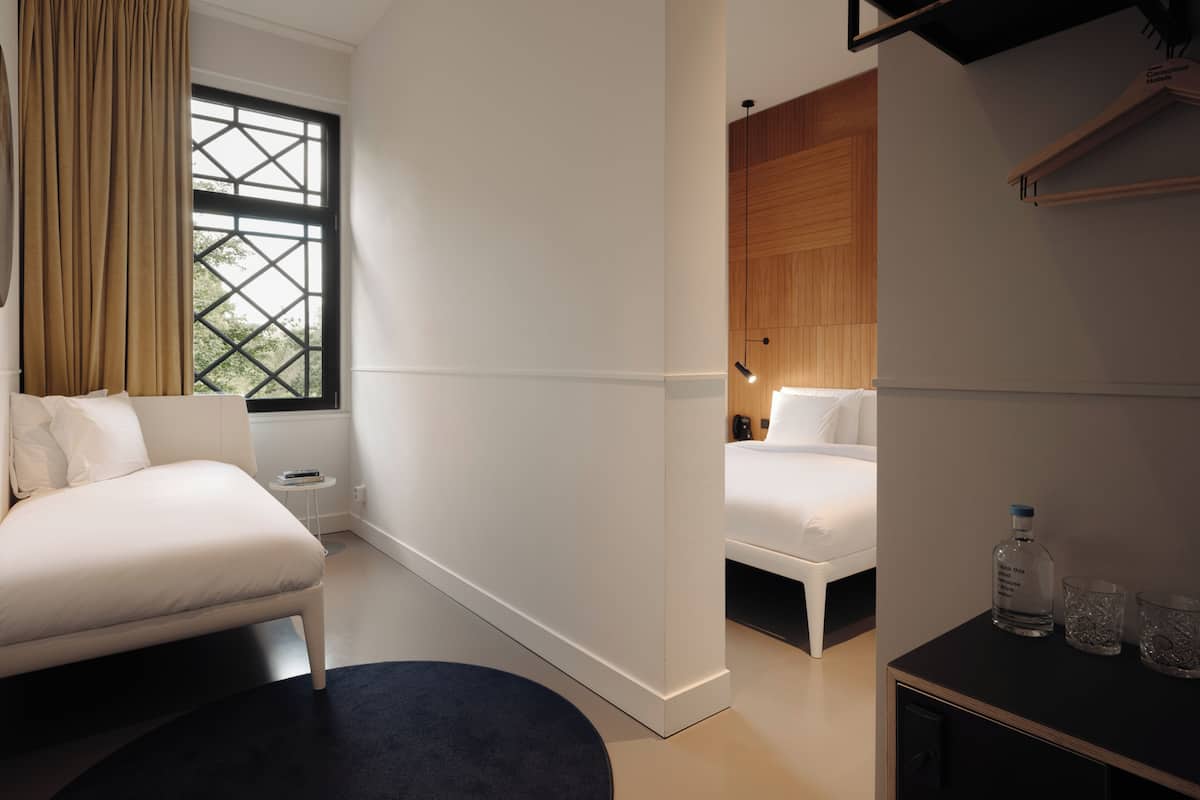 Una habitación de hotel moderna con un ventanal grande, una cama, una mesita con vasos y una botella, y un panel de pared de madera.