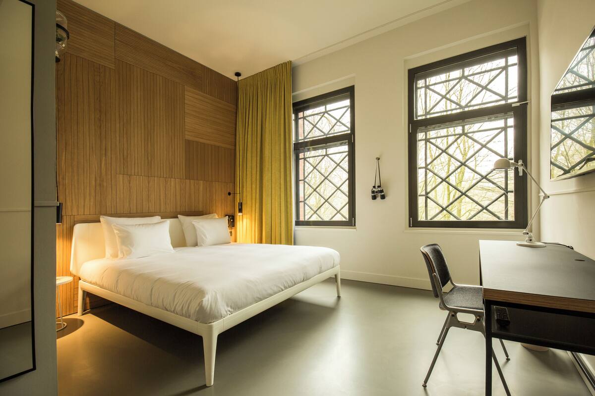 Un dormitorio moderno con cama, escritorio y dos ventanas con refuerzo en forma de cruz.