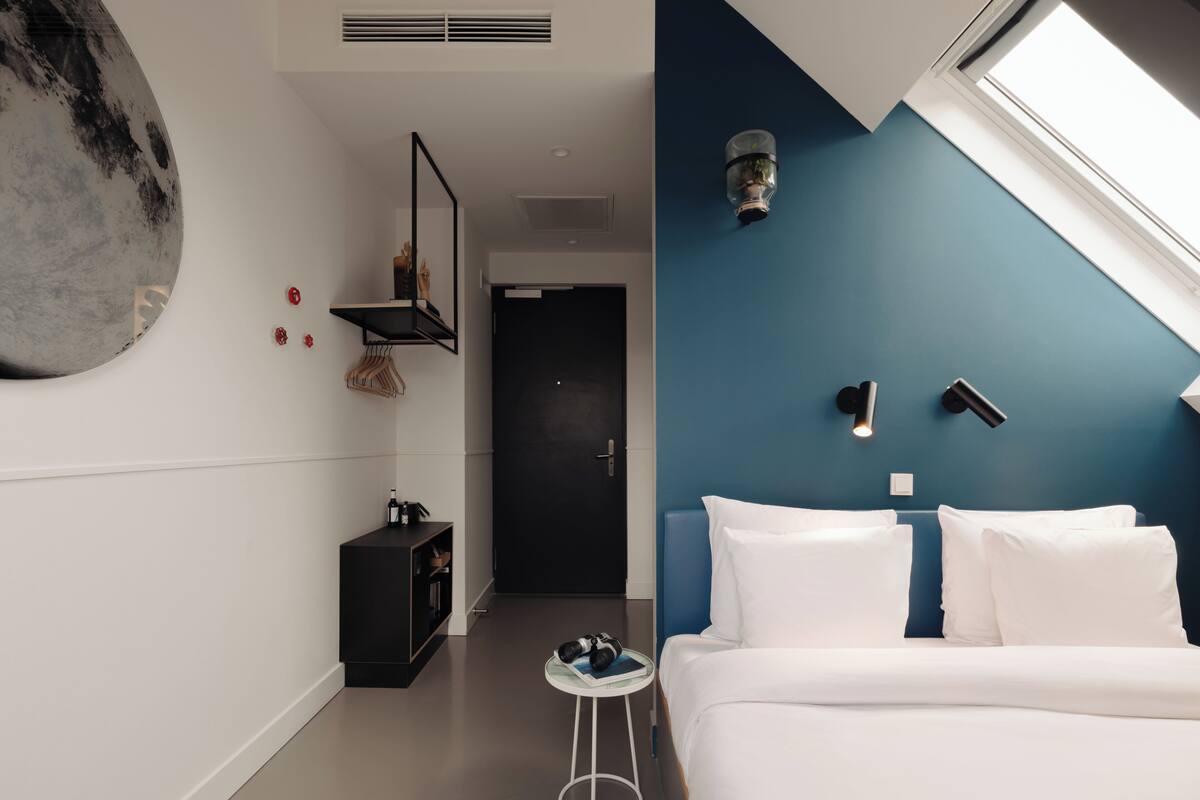 Una habitación de hotel moderna con una cama grande, una mesita auxiliar pequeña, una puerta oscura y una pared azul decorativa.