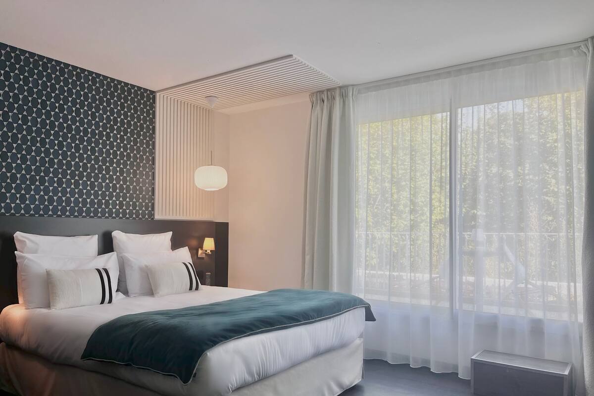 A modern hotel room with a neatly made bed, a large window with sheer curtains, and a patterned wall.