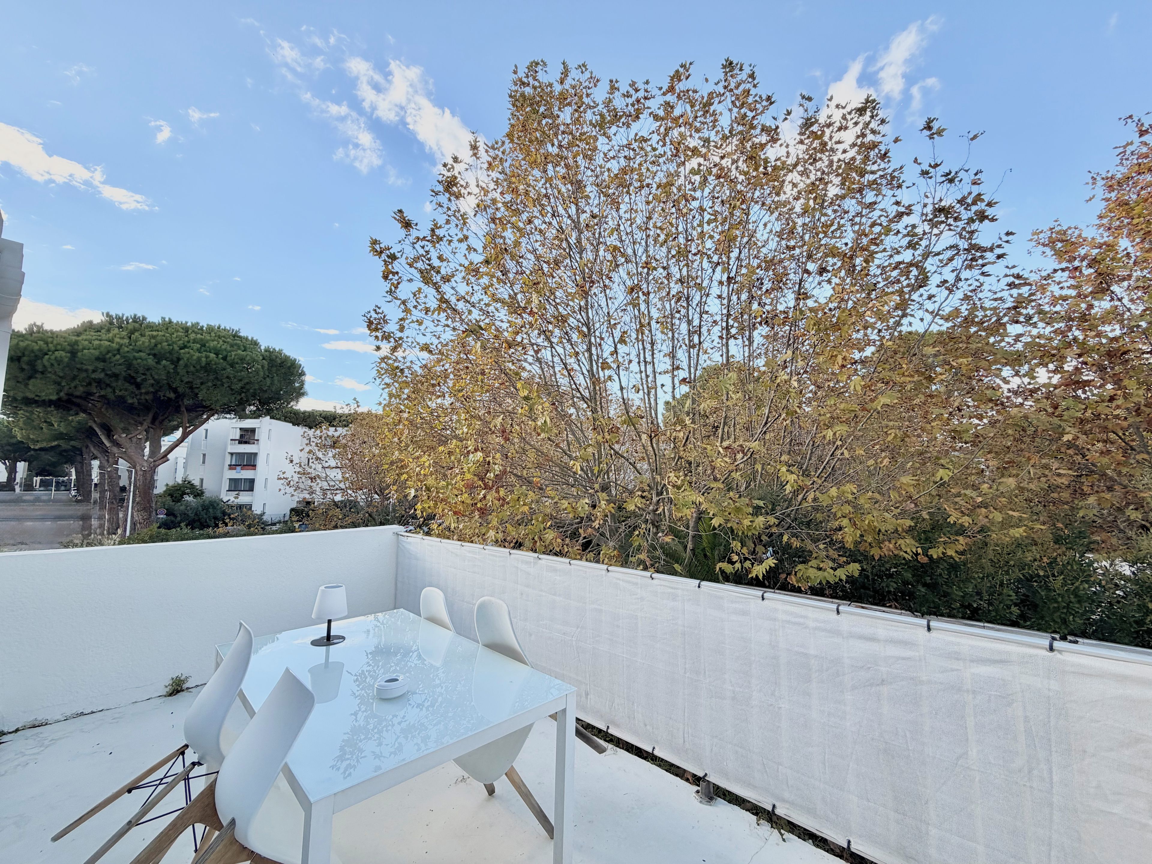 A rooftop terrace with a glass table and chairs, surrounded by trees and a white building.