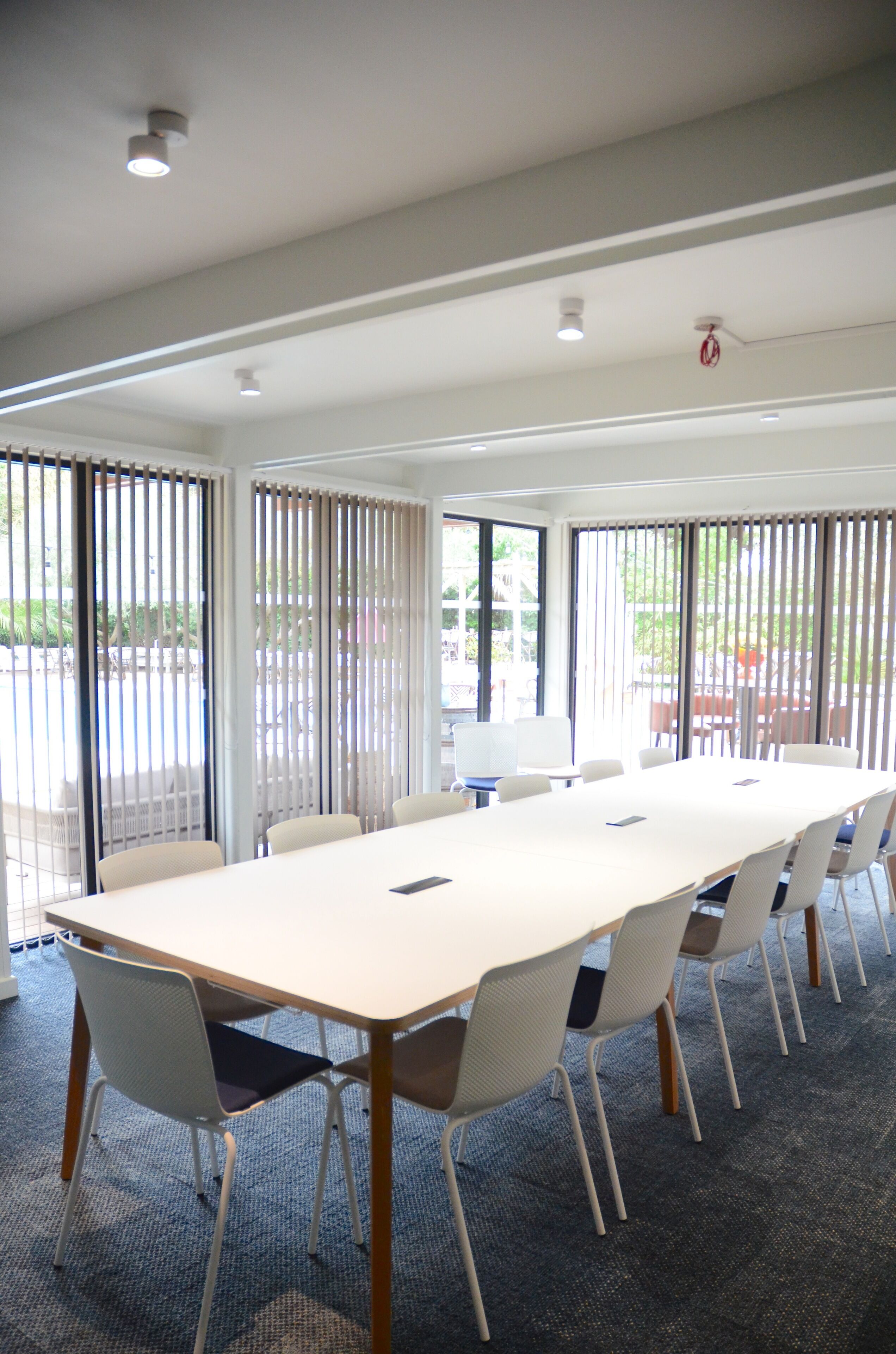Un salón de conferencias con una larga mesa y sillas, ventanas amplias con persianas y piso alfombrado.