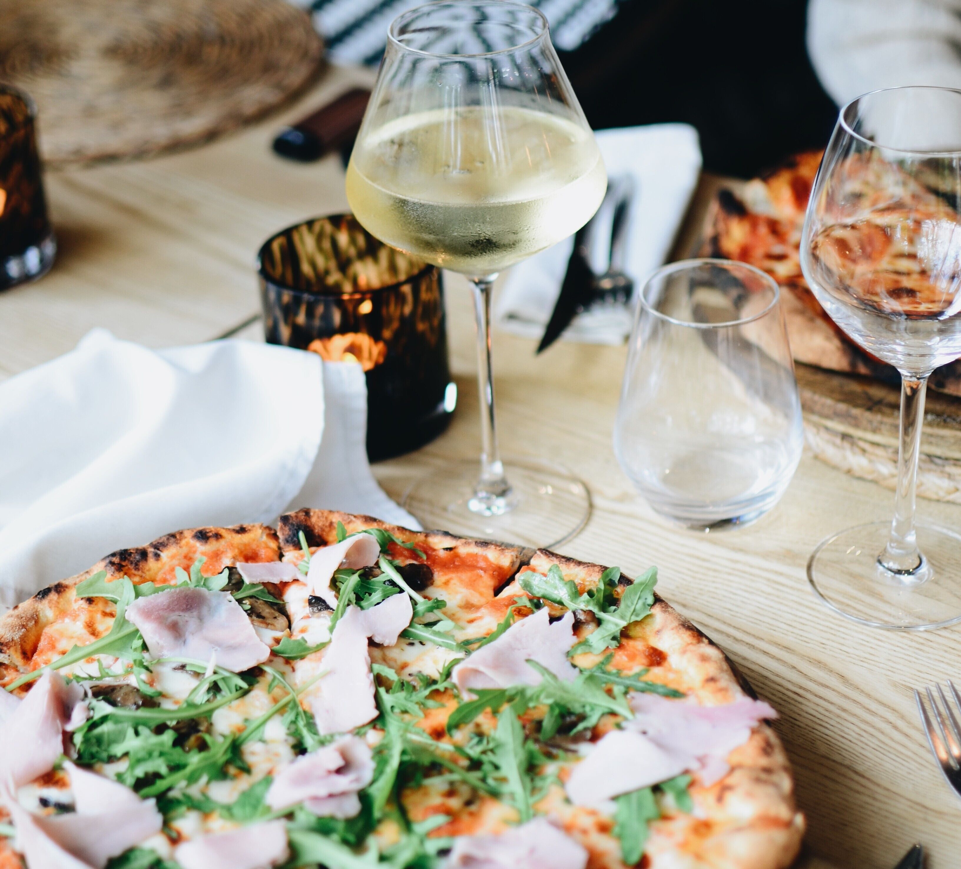 A pizza with greens and ham, a glass of white wine, and a candle on a wooden table.