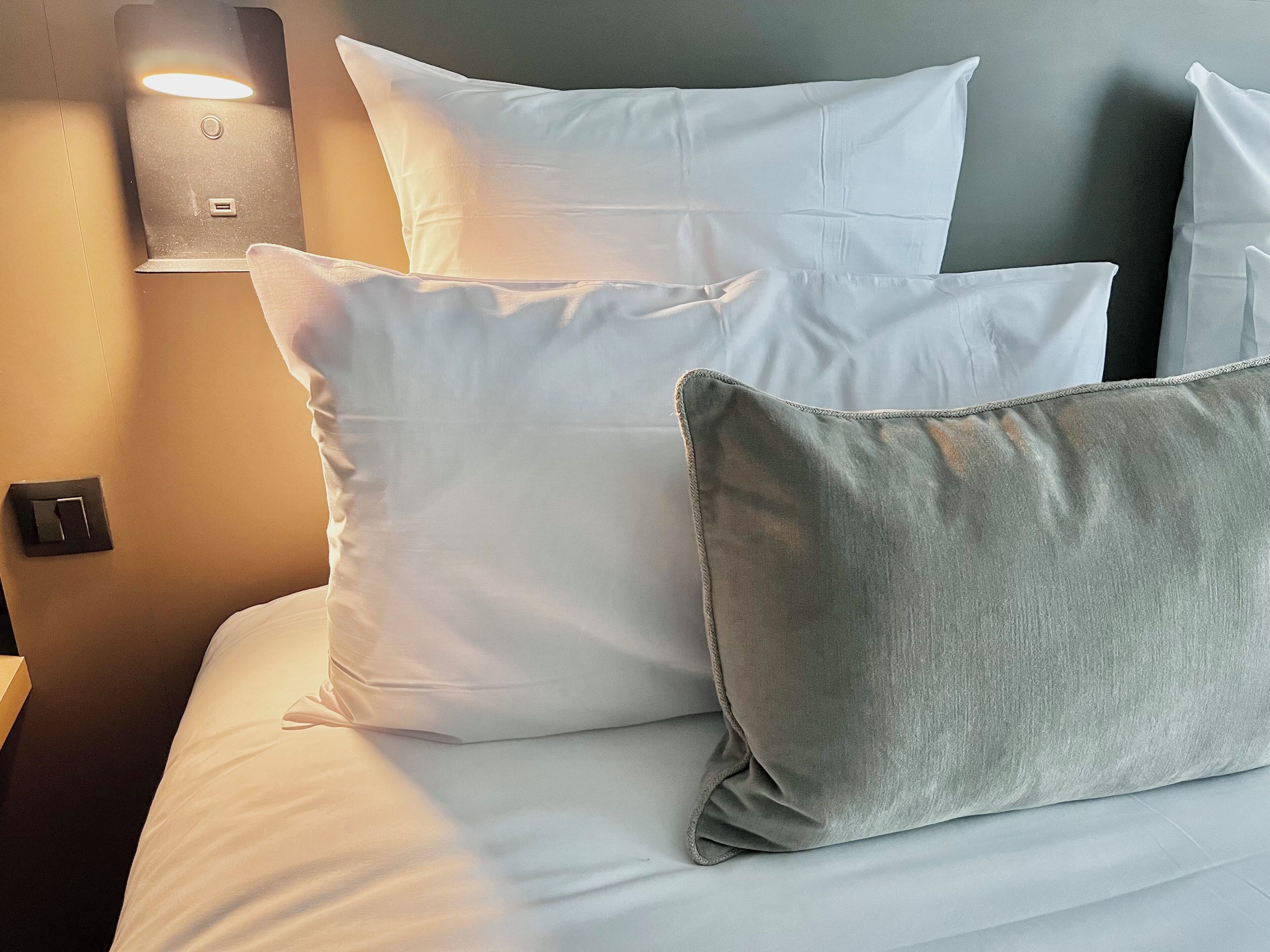 A bed with white and grey pillows, a wall-mounted light, and a power outlet.