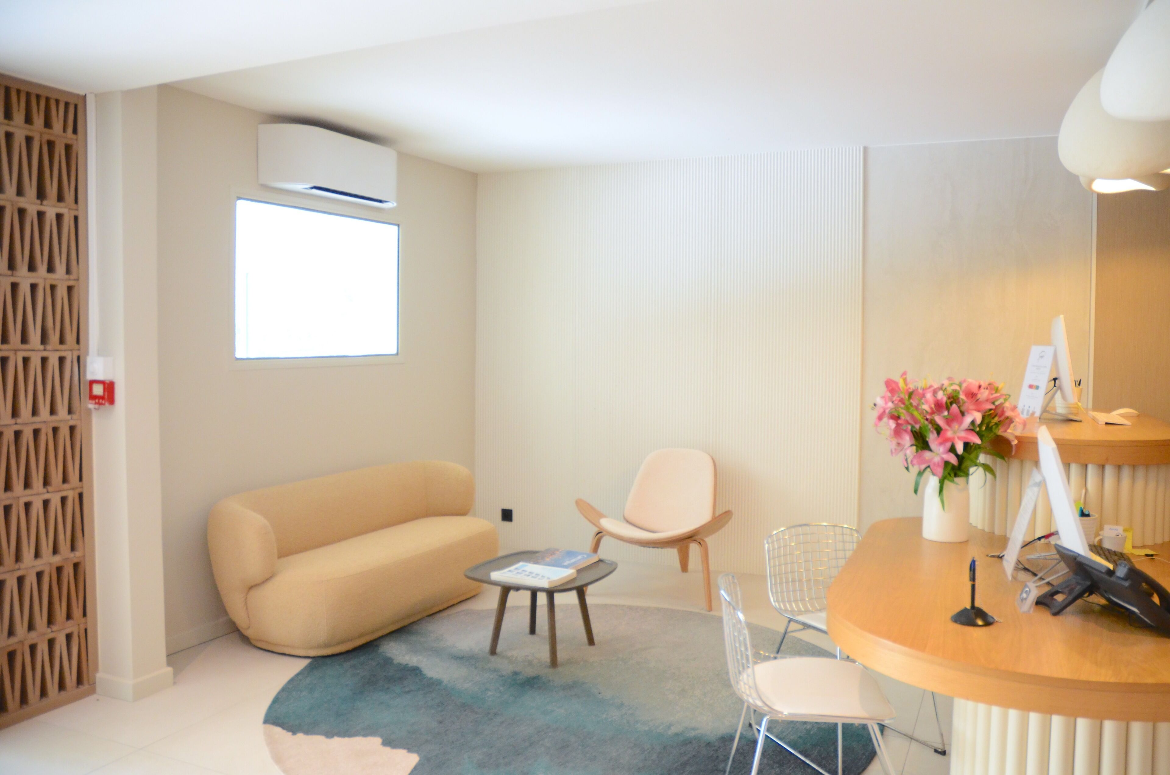 Een moderne hotelkamer met een beige bank, een klein rond tafeltje, een stoel, een bureau met een computer en een vaas met bloemen.